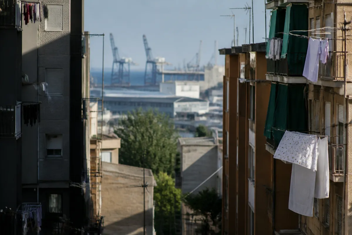 Calle residencial de bloques de los setenta junto a un tramo de costa industrial, luz de media tarde, sin rostros ni rótulos.